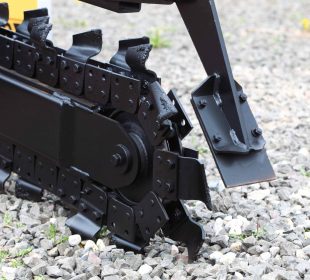 Close-up of a trencher attachment digging into gravel, showing sharp cutting teeth and metal components.