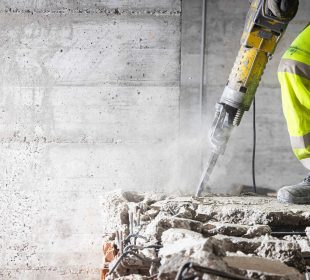 Construction worker in high-visibility gear using a jackhammer to break concrete, with dust, debris, and exposed rebar visible.