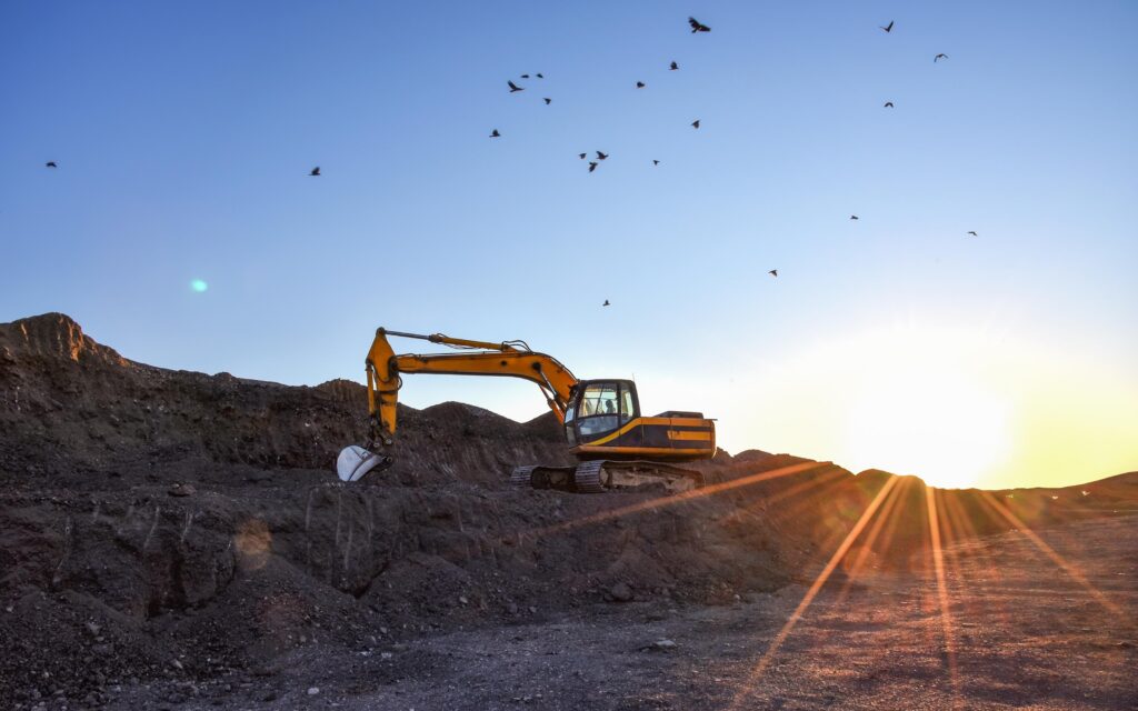 A yellow excavator is parked on a rocky terrain during sunset, with the sun casting long rays across the ground and birds flying in the clear sky above.