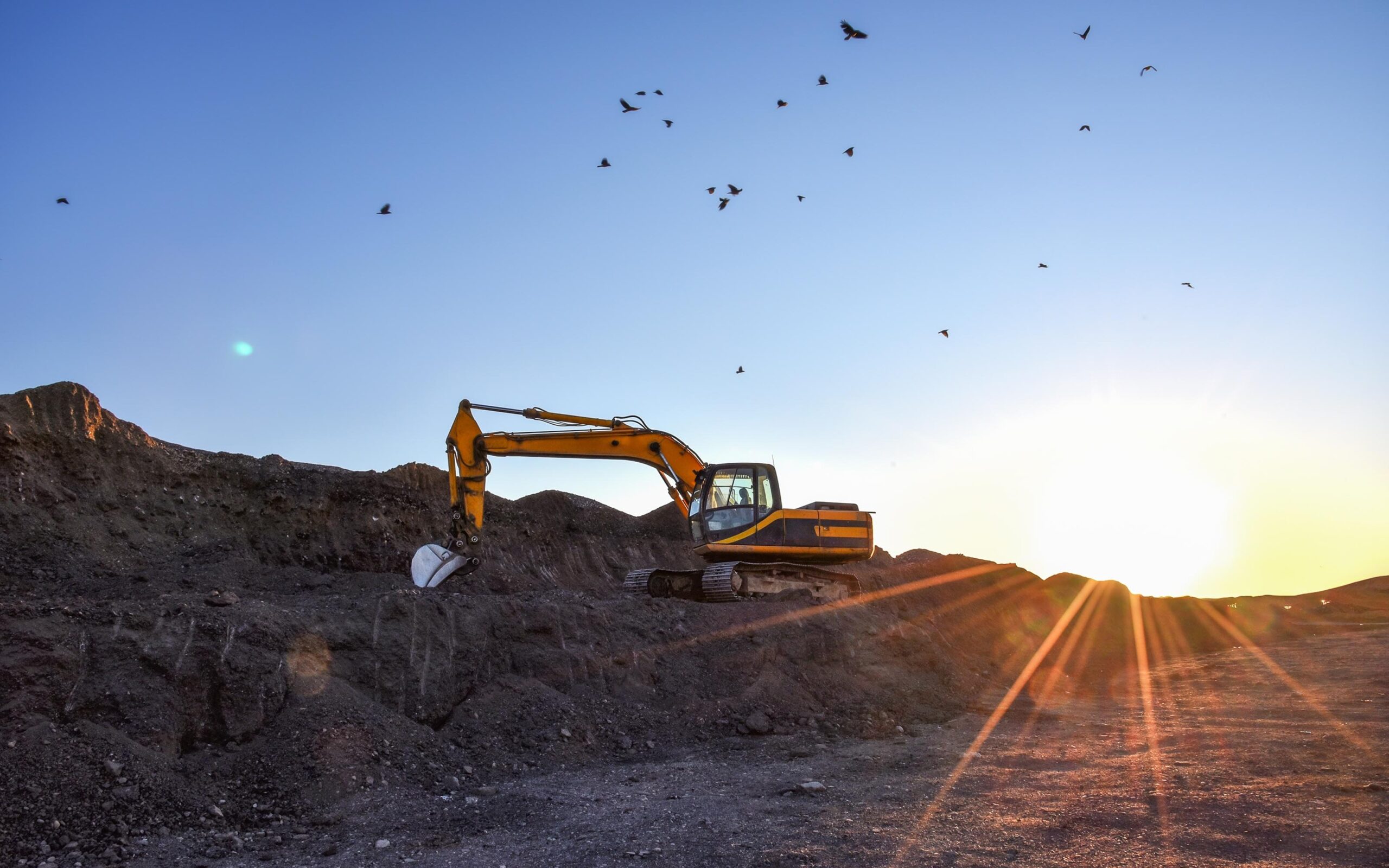 A yellow excavator is parked on a rocky terrain during sunset, with the sun casting long rays across the ground and birds flying in the clear sky above.