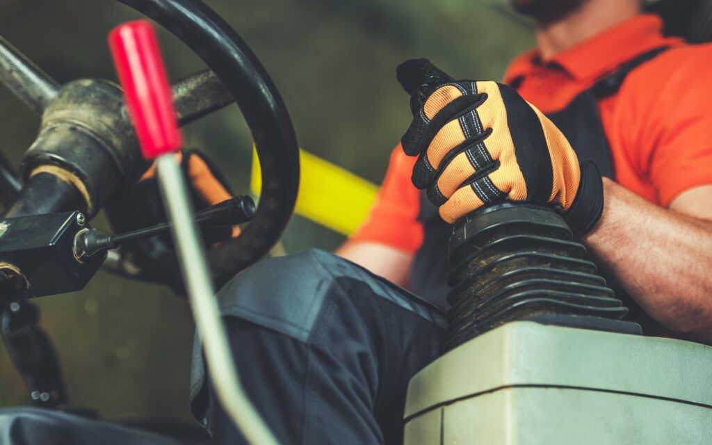 A close-up image of a person operating heavy machinery, focusing on the hands gripping the control lever. The individual is wearing an orange shirt and protective gloves.