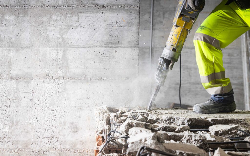 Construction worker in high-visibility gear using a jackhammer to break concrete, with dust, debris, and exposed rebar visible.