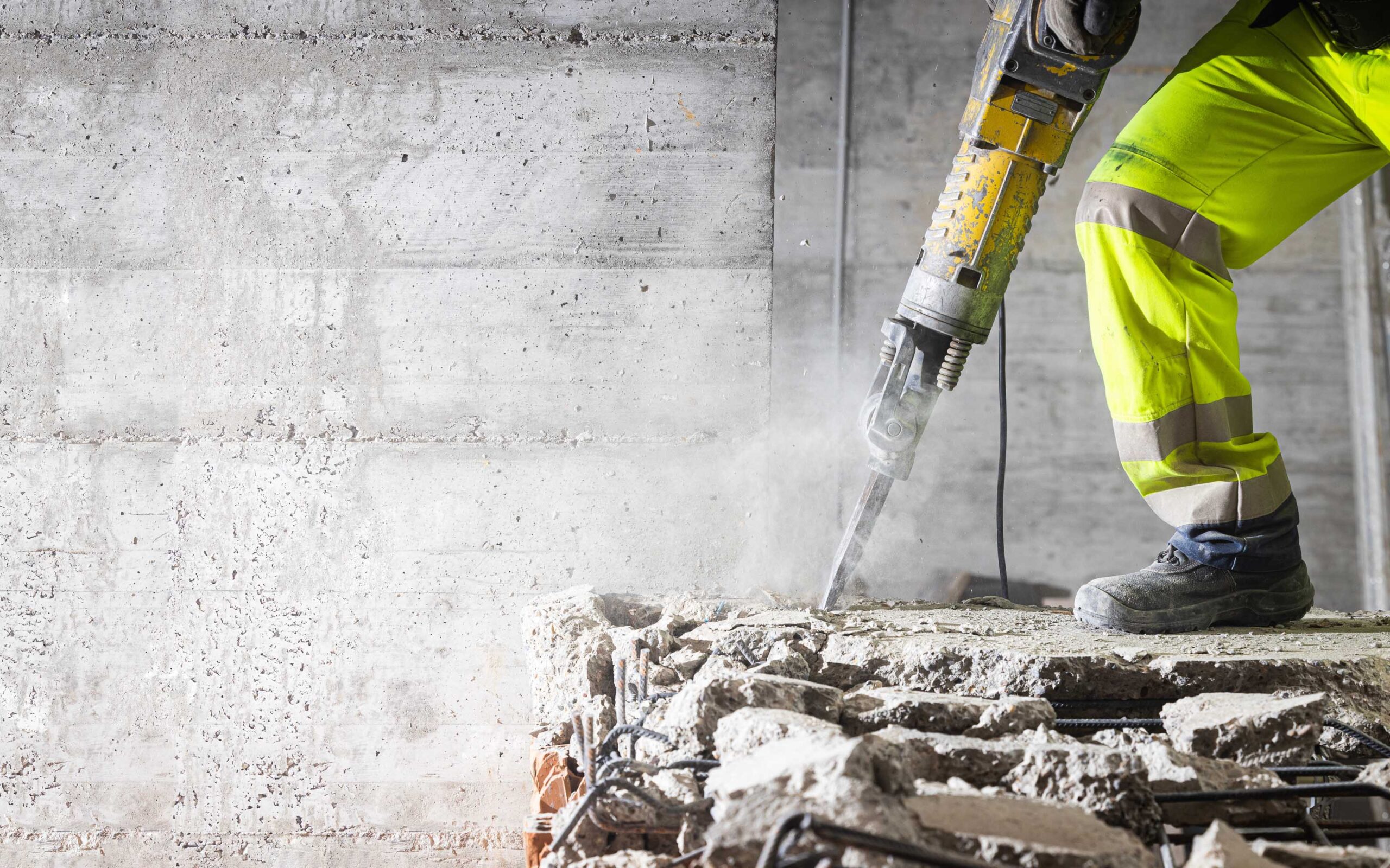 Construction worker in high-visibility gear using a jackhammer to break concrete, with dust, debris, and exposed rebar visible.