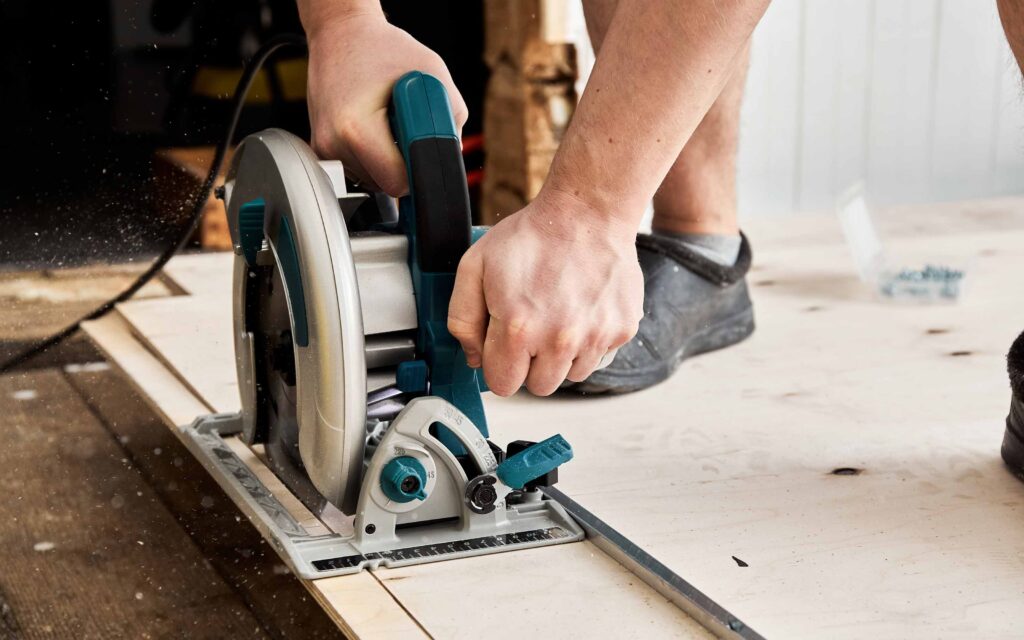 Person using a circular saw with a guide rail to cut a piece of wood on a wooden surface, with tools and screws visible in the background