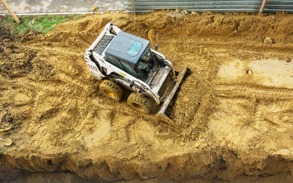 Skid-steer loader moving soil on a construction site, with visible tire tracks, a grassy patch, and a metal barrier in the background.