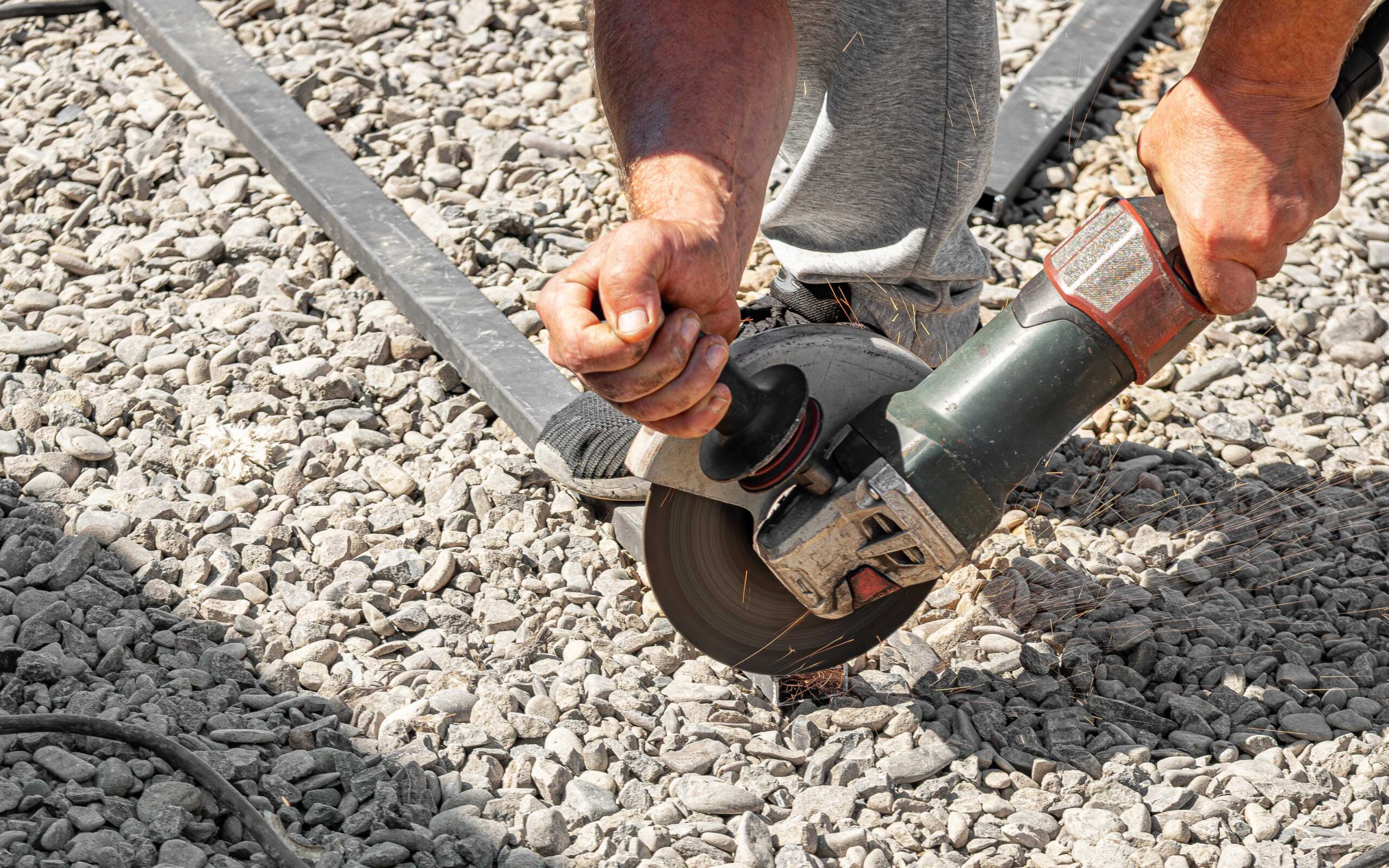 Person using an angle grinder to cut a metal bar, with bright sparks flying and gravel-covered ground beneath.