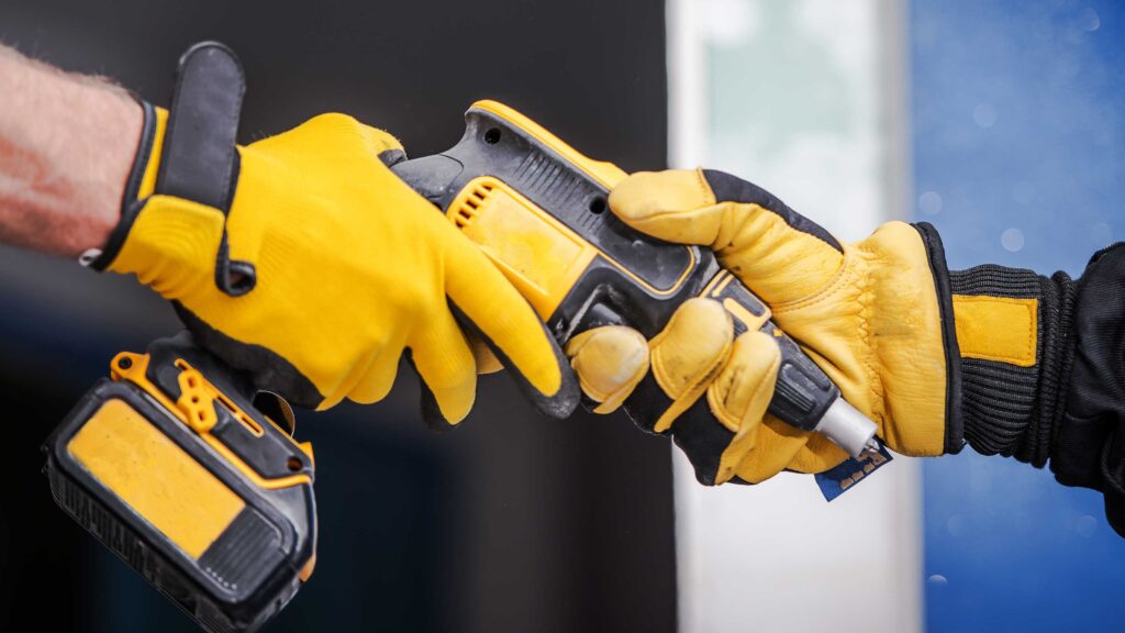 Two workers wearing yellow gloves exchanging a cordless power drill on site.