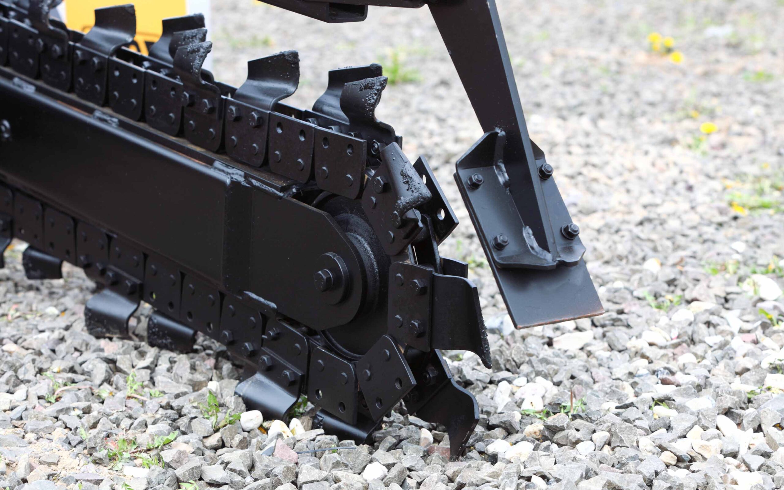 Close-up of a trencher attachment digging into gravel, showing sharp cutting teeth and metal components.