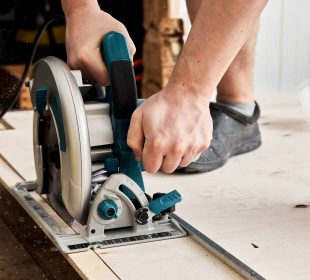 Person using a circular saw with a guide rail to cut a piece of wood on a wooden surface, with tools and screws visible in the background