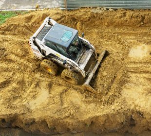 Skid-steer loader moving soil on a construction site, with visible tire tracks, a grassy patch, and a metal barrier in the background.