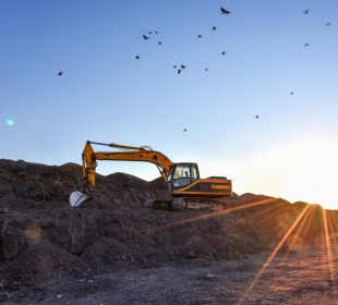 A yellow excavator is parked on a rocky terrain during sunset, with the sun casting long rays across the ground and birds flying in the clear sky above.