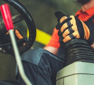 A close-up image of a person operating heavy machinery, focusing on the hands gripping the control lever. The individual is wearing an orange shirt and protective gloves.